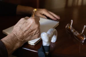 Elderly hand writing in notebook, glasses nearby.