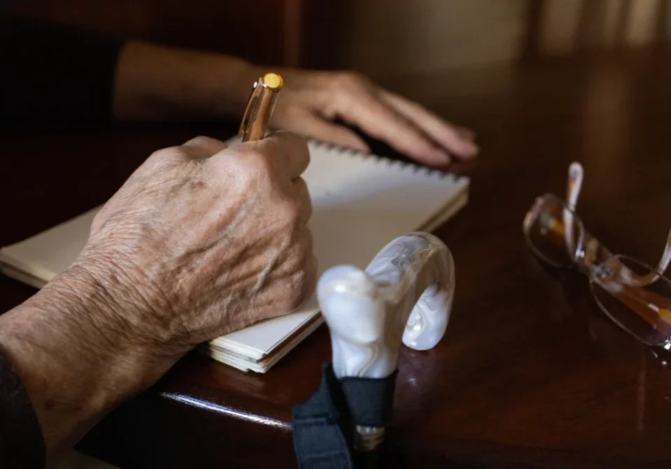 Elderly hand writing in notebook, glasses nearby.